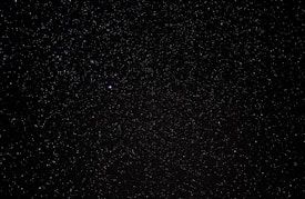 A vast expanse of a night sky filled with countless tiny stars scattered across a deep black background, with a single noticeably brighter star. The scene embodies the vastness of the universe.