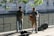 Two musicians perform outdoors beside a canal. One plays a saxophone while wearing a hat and casual attire, and the other plays a guitar in a short-sleeved shirt and shorts. The setting includes a brick building, water, and greenery, suggesting a serene urban environment.