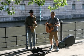 Two musicians perform outdoors beside a canal. One plays a saxophone while wearing a hat and casual attire, and the other plays a guitar in a short-sleeved shirt and shorts. The setting includes a brick building, water, and greenery, suggesting a serene urban environment.