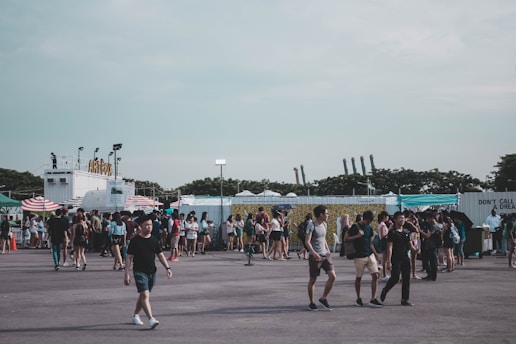 A large outdoor festival or market with people walking and gathering around various stalls and food stands. There are several tents and a structure with the word 'Artbox' visible. The atmosphere seems lively with a diverse crowd enjoying the event. The sky is overcast, and the area is surrounded by greenery and industrial elements in the background.