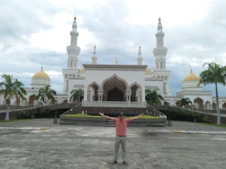 A man is standing with outstretched arms in front of an ornate mosque featuring white walls and golden domes. The structure is surrounded by palm trees and set against a cloudy sky.