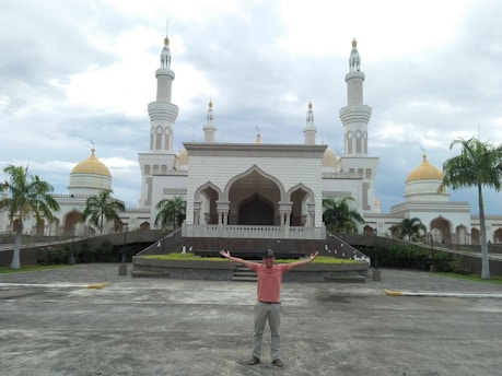 A man is standing with outstretched arms in front of an ornate mosque featuring white walls and golden domes. The structure is surrounded by palm trees and set against a cloudy sky.