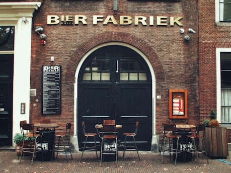 A brick building facade features a large black door with an arched top, flanked by a sign displaying text. Above the door, large gold letters spell out 'BIER FABRIEK.' Below, round tables and stools are arranged, creating an outdoor seating area. There are potted plants and a small menu board on the wall.