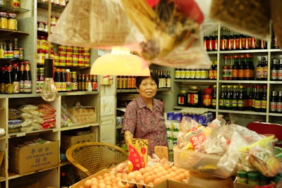 A woman stands behind a counter inside a small, traditional grocery store. The shelves are stocked with various bottled sauces, seasonings, and packaged goods. There are several bags filled with spices and dried goods hanging from the ceiling. A warm light hangs above, illuminating a basket of eggs on the counter.