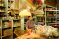 A woman stands behind a counter inside a small, traditional grocery store. The shelves are stocked with various bottled sauces, seasonings, and packaged goods. There are several bags filled with spices and dried goods hanging from the ceiling. A warm light hangs above, illuminating a basket of eggs on the counter.
