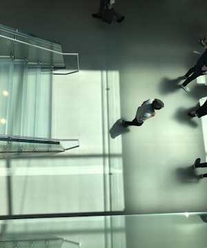 An overhead view of a minimalist indoor space featuring a glass staircase and several people walking across a large tiled floor. Sunlight casts long, sharp shadows and light patterns across the floor, giving the scene a dynamic effect.