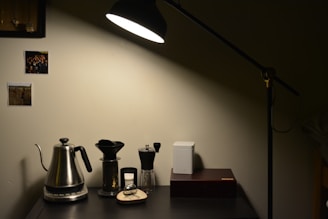 A cozy and minimalist home setup with a black desk lamp illuminating a section of a dark desk. On the desk are a metallic kettle, coffee dripper, manual coffee grinder, and a closed white container next to a rectangular box. The wall behind features two pinned photographs, adding a personal touch. The lighting creates a warm and inviting atmosphere.