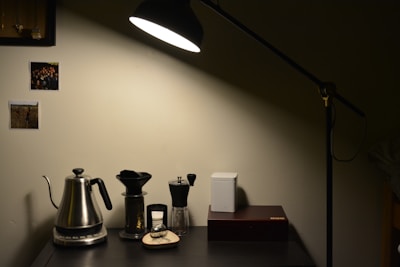 A cozy and minimalist home setup with a black desk lamp illuminating a section of a dark desk. On the desk are a metallic kettle, coffee dripper, manual coffee grinder, and a closed white container next to a rectangular box. The wall behind features two pinned photographs, adding a personal touch. The lighting creates a warm and inviting atmosphere.