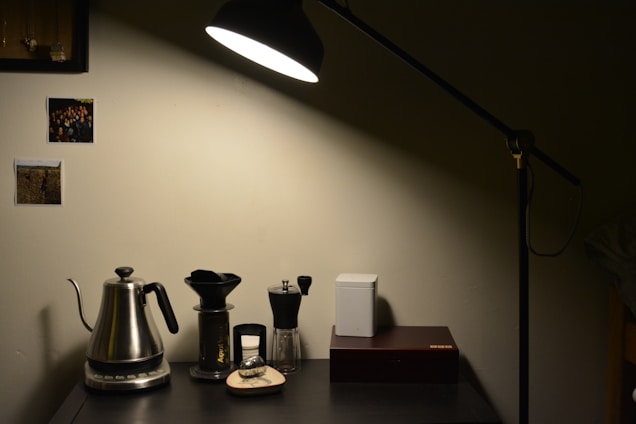 A cozy and minimalist home setup with a black desk lamp illuminating a section of a dark desk. On the desk are a metallic kettle, coffee dripper, manual coffee grinder, and a closed white container next to a rectangular box. The wall behind features two pinned photographs, adding a personal touch. The lighting creates a warm and inviting atmosphere.