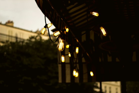 A string of vintage Edison-style bulbs is hanging under a roof awning, casting a warm, ambient glow. The background shows out-of-focus foliage and the outline of buildings, hinting at an outdoor setting.