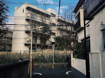 A residential area with multiple apartment buildings. There are several electrical wires overhead, and a streetlamp is visible. The scene on the left shows a woman in a face mask and the right shows someone riding a bicycle, suggesting everyday urban life. Trees and tall grasses are situated behind a green fence, adding a touch of nature to the urban environment.