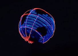 A globe-like structure illuminated with vibrant neon lights is set against a dark background. The structure features interlacing blue and red lights in swirling patterns, creating an impression of an abstract representation of Earth.