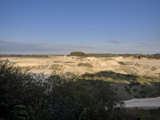 A vast open landscape with large earth mounds and exposed soil, surrounded by greenery. The land appears to be used for mining or quarrying activities, with varying levels of elevation and visible paths or tracks carved into the terrain. In the distance, a line of trees marks the horizon under a clear blue sky.