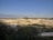 A vast open landscape with large earth mounds and exposed soil, surrounded by greenery. The land appears to be used for mining or quarrying activities, with varying levels of elevation and visible paths or tracks carved into the terrain. In the distance, a line of trees marks the horizon under a clear blue sky.