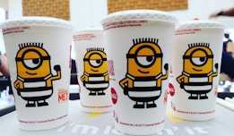 Four white beverage cups with illustrations of a yellow cartoon character wearing a striped outfit and goggles are placed closely together on a table. The cups have branding for 'Despicable Me 3' and are marked with a Coca-Cola logo. The background is blurred, with hints of a room and people.
