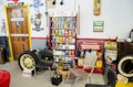 A vintage garage scene with various retro automotive signs and memorabilia. There are several cans of old car products displayed on a shelf, including oil and additives. A large yellow car wheel and tire lean against a wall. Multiple old-fashioned chairs are scattered around the room. The walls are decorated with vintage advertisements and signs for brands like Chevrolet, Firestone, and Quaker State.