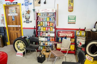 A vintage garage scene with various retro automotive signs and memorabilia. There are several cans of old car products displayed on a shelf, including oil and additives. A large yellow car wheel and tire lean against a wall. Multiple old-fashioned chairs are scattered around the room. The walls are decorated with vintage advertisements and signs for brands like Chevrolet, Firestone, and Quaker State.