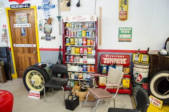 A vintage garage scene with various retro automotive signs and memorabilia. There are several cans of old car products displayed on a shelf, including oil and additives. A large yellow car wheel and tire lean against a wall. Multiple old-fashioned chairs are scattered around the room. The walls are decorated with vintage advertisements and signs for brands like Chevrolet, Firestone, and Quaker State.