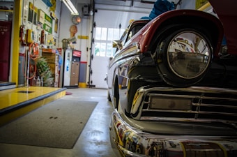 A vintage car is parked inside a garage with various tools and retro decorations on the walls. The car's shiny surface reflects its surroundings, including colorful signs and equipment. The garage has a slightly cluttered yet organized feel, with hoses, beverage machines, and industrial decor visible.