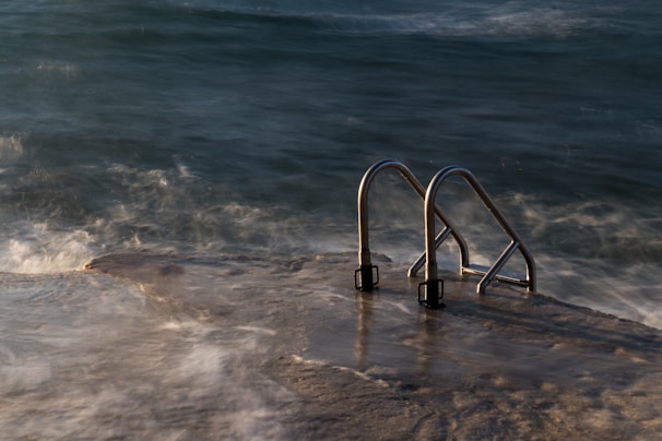 Stainless steel handrails leading into the ocean, with waves gently crashing against a concrete platform. The scene is captured during twilight, adding a mysterious and serene ambiance.