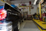 A vintage garage filled with classic decor and automotive memorabilia. An old black car with a shiny finish, possibly from the 1950s, is parked inside. The walls are adorned with retro signs and advertisements, such as 'MOTOR OIL' and other automotive brands. Shelves stocked with antique oil cans and various car parts contribute to the nostalgic vibe. A workbench with tools and equipment can be seen, and there’s a collection of model cars displayed above. The checkered pattern on the ramp adds to the retro theme.