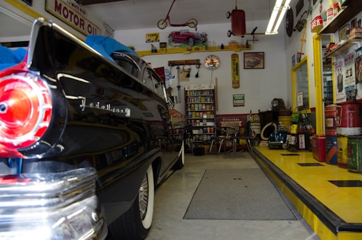 A vintage garage filled with classic decor and automotive memorabilia. An old black car with a shiny finish, possibly from the 1950s, is parked inside. The walls are adorned with retro signs and advertisements, such as 'MOTOR OIL' and other automotive brands. Shelves stocked with antique oil cans and various car parts contribute to the nostalgic vibe. A workbench with tools and equipment can be seen, and there’s a collection of model cars displayed above. The checkered pattern on the ramp adds to the retro theme.