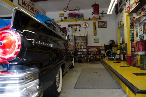 A vintage garage filled with classic decor and automotive memorabilia. An old black car with a shiny finish, possibly from the 1950s, is parked inside. The walls are adorned with retro signs and advertisements, such as 'MOTOR OIL' and other automotive brands. Shelves stocked with antique oil cans and various car parts contribute to the nostalgic vibe. A workbench with tools and equipment can be seen, and there’s a collection of model cars displayed above. The checkered pattern on the ramp adds to the retro theme.