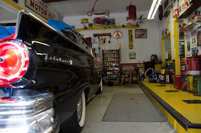 A vintage garage filled with classic decor and automotive memorabilia. An old black car with a shiny finish, possibly from the 1950s, is parked inside. The walls are adorned with retro signs and advertisements, such as 'MOTOR OIL' and other automotive brands. Shelves stocked with antique oil cans and various car parts contribute to the nostalgic vibe. A workbench with tools and equipment can be seen, and there’s a collection of model cars displayed above. The checkered pattern on the ramp adds to the retro theme.