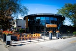 A modern, circular restaurant with large glass windows and a patio featuring several orange chairs and black tables. The establishment is surrounded by greenery, including large trees and vibrant flower pots at the entrance. The sign on the building displays the name Culaccino in bold letters, and the ambiance suggests an inviting outdoor dining area bathed in bright sunlight.