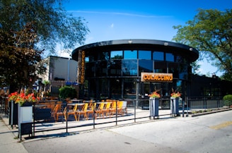 A modern, circular restaurant with large glass windows and a patio featuring several orange chairs and black tables. The establishment is surrounded by greenery, including large trees and vibrant flower pots at the entrance. The sign on the building displays the name Culaccino in bold letters, and the ambiance suggests an inviting outdoor dining area bathed in bright sunlight.