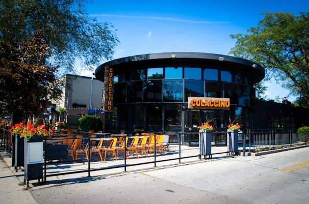 A modern, circular restaurant with large glass windows and a patio featuring several orange chairs and black tables. The establishment is surrounded by greenery, including large trees and vibrant flower pots at the entrance. The sign on the building displays the name Culaccino in bold letters, and the ambiance suggests an inviting outdoor dining area bathed in bright sunlight.