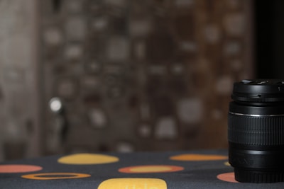 A camera lens is placed on a surface with a dark cloth featuring large colored circles. The background is blurred, showcasing geometric and somewhat abstract patterns.