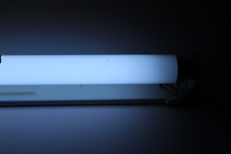 A fluorescent light fixture with a tubular bulb is mounted on a wall. The bulb emits a soft blue-white glow, illuminating the dark surrounding area. The fixture itself appears to be metallic and minimalistic in design.