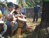 A woman and a young child are joyfully playing drums together outdoors. They are sitting on a large piece of wood in a garden or park setting. The child is enthusiastically hitting the drum, and the woman appears supportive, sharing in the moment. There are other people in the background, and the setting is sunny with greenery around.