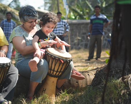 A woman and a young child are joyfully playing drums together outdoors. They are sitting on a large piece of wood in a garden or park setting. The child is enthusiastically hitting the drum, and the woman appears supportive, sharing in the moment. There are other people in the background, and the setting is sunny with greenery around.