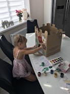 A young child is seated at a dining table, engaged in an art project. She is painting on a large cardboard box with various colors. The table is scattered with watercolor paints, a palette, and sheets of paper with drawings. In the background, there is a window with a roller blind, a vase with flowers, and decorative elephant figurines.
