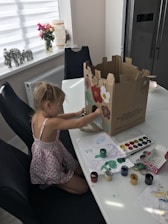 A young child is seated at a dining table, engaged in an art project. She is painting on a large cardboard box with various colors. The table is scattered with watercolor paints, a palette, and sheets of paper with drawings. In the background, there is a window with a roller blind, a vase with flowers, and decorative elephant figurines.