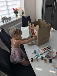 A young child is seated at a dining table, engaged in an art project. She is painting on a large cardboard box with various colors. The table is scattered with watercolor paints, a palette, and sheets of paper with drawings. In the background, there is a window with a roller blind, a vase with flowers, and decorative elephant figurines.