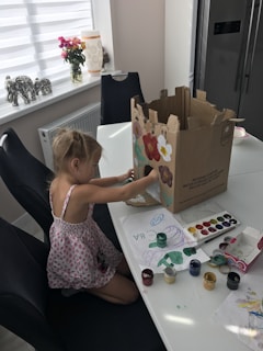 A young child is seated at a dining table, engaged in an art project. She is painting on a large cardboard box with various colors. The table is scattered with watercolor paints, a palette, and sheets of paper with drawings. In the background, there is a window with a roller blind, a vase with flowers, and decorative elephant figurines.