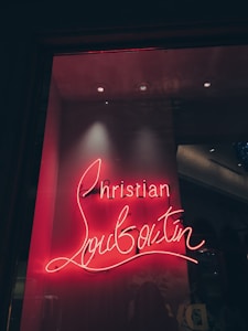 A glowing neon sign displaying the name 'Christian Louboutin' in a cursive font. The sign is set against a dark background, giving it a vibrant contrast. The lighting creates a warm, inviting ambiance.