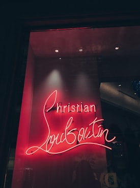 A glowing neon sign displaying the name 'Christian Louboutin' in a cursive font. The sign is set against a dark background, giving it a vibrant contrast. The lighting creates a warm, inviting ambiance.
