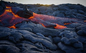 Molten lava flows over a dark, rocky surface, radiating bright orange and red hues in striking contrast to the cooling, blackened stone. The texture of the rocks is rough and uneven, with the lava's movement appearing fluid and dynamic.
