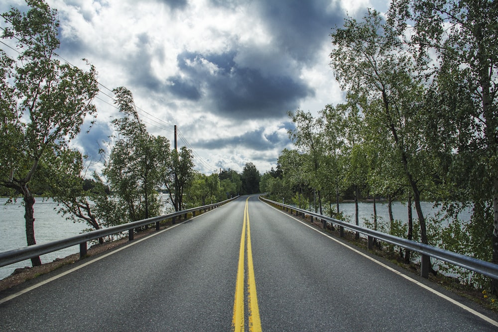 gray concrete road between green trees under white clouds and blue sky during daytime