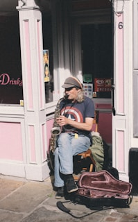 An individual sitting on a wooden chair is playing a saxophone on a sunny street corner. The person is wearing a cap and a t-shirt featuring a large star emblem. The background consists of a pink-painted shop facade with various signs and a partially opened door. There is a saxophone case on the ground next to the musician.