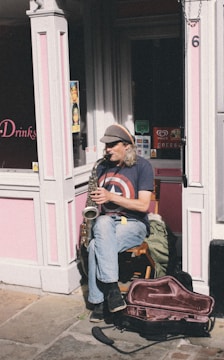 An individual sitting on a wooden chair is playing a saxophone on a sunny street corner. The person is wearing a cap and a t-shirt featuring a large star emblem. The background consists of a pink-painted shop facade with various signs and a partially opened door. There is a saxophone case on the ground next to the musician.