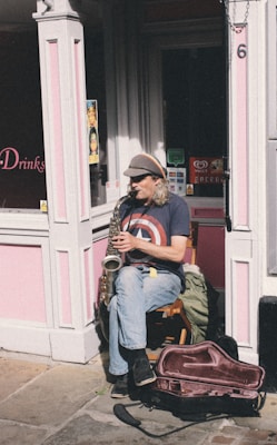 An individual sitting on a wooden chair is playing a saxophone on a sunny street corner. The person is wearing a cap and a t-shirt featuring a large star emblem. The background consists of a pink-painted shop facade with various signs and a partially opened door. There is a saxophone case on the ground next to the musician.