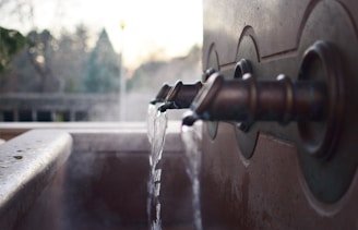 Water flows from three vintage-style metal faucets mounted on a stone wall into a basin. Steam rises in the cool air, suggesting warm water flow. In the blurred background, outdoor greenery and faint structures are visible.