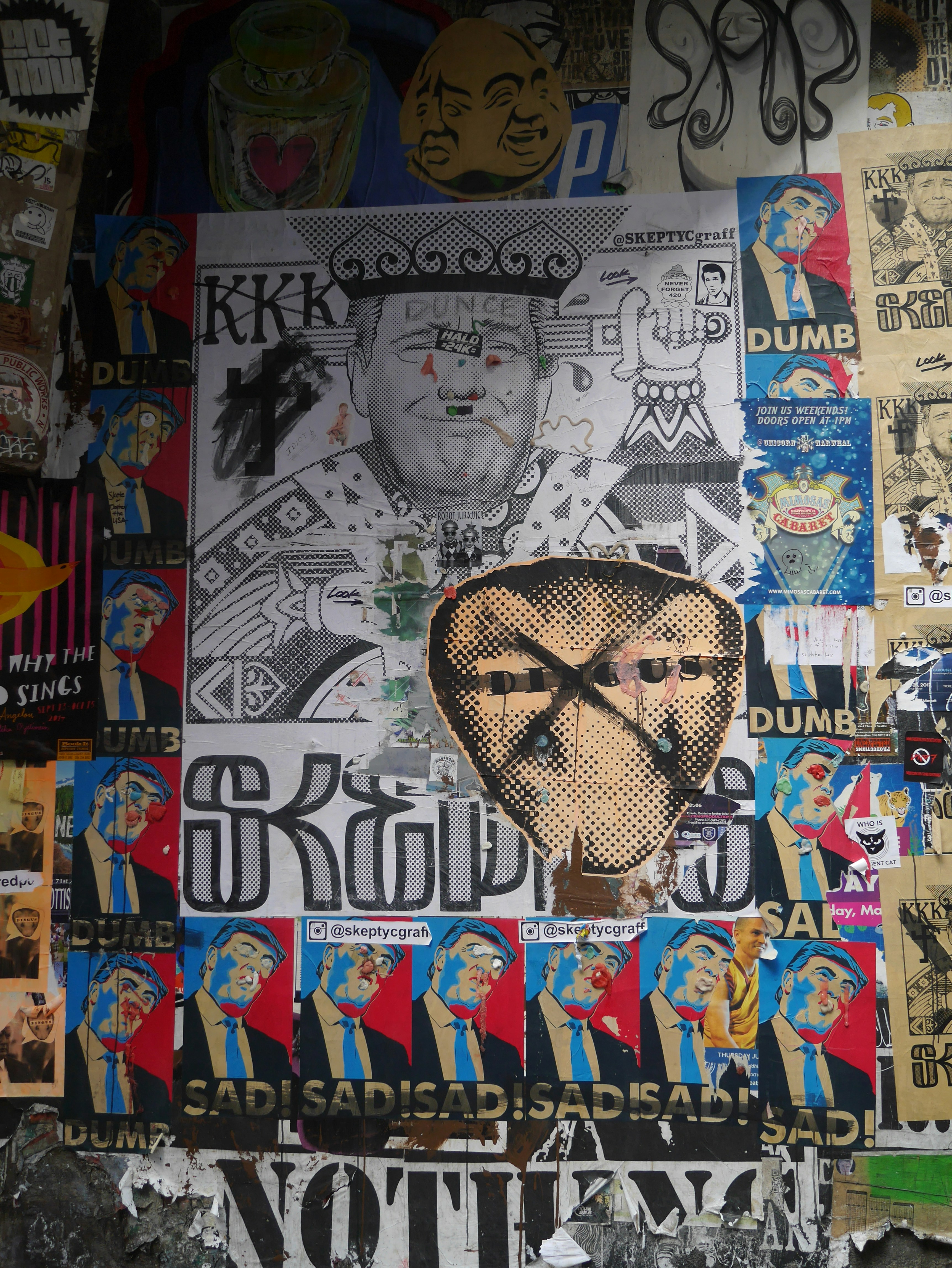 A collage of various street art posters, featuring political and satirical elements. Central to the composition is a large monochrome poster of a man with 'Dunce' and 'KKK' written on it, surrounded by smaller colorful images depicting the same figure with expressions of discontent and words like 'Dumb' and 'Sad'. Other posters in the mix include abstract designs and images of different faces.