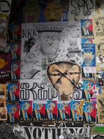A collage of various street art posters, featuring political and satirical elements. Central to the composition is a large monochrome poster of a man with 'Dunce' and 'KKK' written on it, surrounded by smaller colorful images depicting the same figure with expressions of discontent and words like 'Dumb' and 'Sad'. Other posters in the mix include abstract designs and images of different faces.