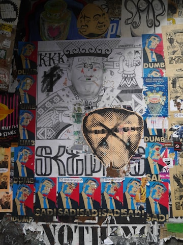 A collage of various street art posters, featuring political and satirical elements. Central to the composition is a large monochrome poster of a man with 'Dunce' and 'KKK' written on it, surrounded by smaller colorful images depicting the same figure with expressions of discontent and words like 'Dumb' and 'Sad'. Other posters in the mix include abstract designs and images of different faces.